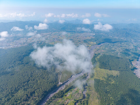 Aerial view of the Sumberurip area featuring dense green forests, a winding river valley, and scattered clouds over the mountainous landscape of Pronojiwo, East Java, Indonesia.