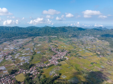 Aerial view of the village of Sumberurip nestled among lush green rice paddies and agricultural fields in the mountainous region of Pronojiwo, East Java, Indonesia.