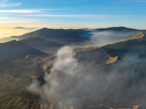 Aerial view of Mount Bromo at sunrise with volcanic smoke rising from the crater, rugged terrain, and golden light illuminating the surrounding mountains in Tosari, East Java, Indonesia.