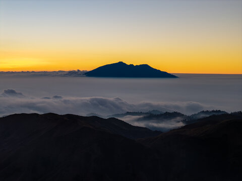 Aerial view of a mountain peak rising above a dense sea of clouds during a golden sunrise in the highlands of East Java, Kecamatan Tosari, Jawa Timur, Indonesia.