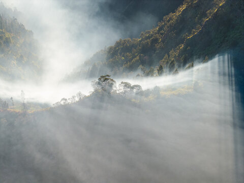 Aerial view of misty mountain slopes near Sukapura, featuring dramatic sunbeams piercing through morning fog, lush green trees, and rugged terrain in Sukapura, East Java, Indonesia.