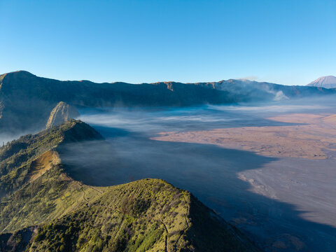 Aerial view of the volcanic landscape of Mount Bromo with mist rolling over the vast caldera and rugged mountain ridges under a clear blue sky in Sukapura, East Java, Indonesia.