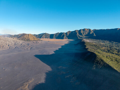 Aerial view of Mount Bromo volcanic landscape with the vast sand sea, rugged crater rims, and dramatic shadows under a clear blue sky in Sukapura, East Java, Indonesia.