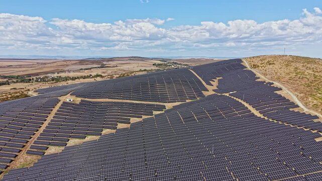 A vast field of solar panels stretches across a hillside in a rural area. The sun shines on the panels as they absorb sunlight. Clouds are visible in the sky.