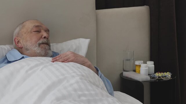 Senior male patient sleeping in retirement home bed with pills on bedside table