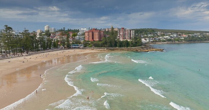 Sydney, Manly Beach coastline with turquoise water, people relax, swim and sunbath on golden sand, beachfront buildings and esplanade under bright blue sky, ideal seaside scene. Aerial view flight