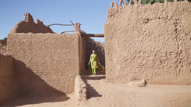 Cinematic tracking shot of an elegant woman wearing a vibrant green embroidered outfit walking between traditional mud brick walls in AlUla