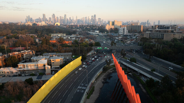 Melbourne, Victoria, Australia - 18 August 2025: Aerial view of Moonee Ponds Creek, city skyline, highway traffic, yellow sound wall, and orange trail art installation.
