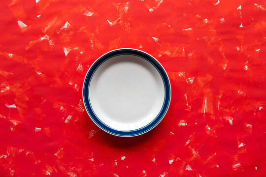 White plate with blue rim sits atop textured red surface. Bright colors create bold, minimalist composition. Perfect for food, dining, or cooking branding