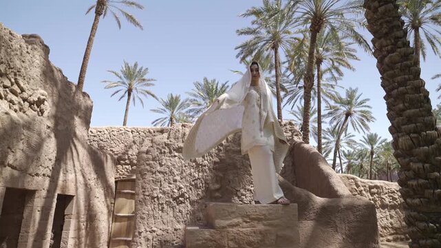 Cinematic low angle shot of an elegant woman in a white embroidered dress and flowing shawl standing on mud brick ruins in AlUla palm oasis