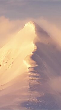 Snowy mountain peak illuminated by golden sunrise light with blowing snow clouds video footage