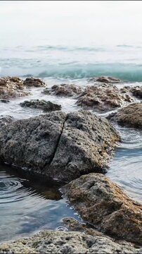 Ocean waves crashing gently against wet rocky shoreline rocks during daytime vertical stock footage clip