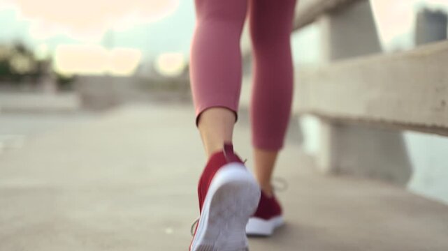 Running shoes with red laces stand on pavement beside a waterfront path. The running shoes fill the frame above athletic feet, with crossed shoelaces, a metal railing, and water beyond the path.