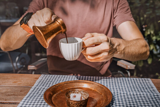 Man pouring Turkish coffee from copper cezve into white cup at wooden cafe table with checkered cloth and copper tray.
