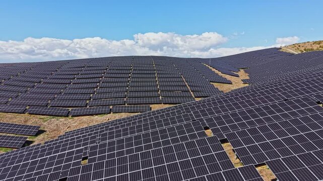 Rows of solar panels stretch across a hilly landscape under a clear blue sky. The scene shows renewable energy being harnessed in an open field.