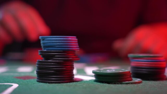 Black poker chips stack tall on green felt. Red glow lights the scene softly. Blurred hands imply concentration. Tension hangs in the gambling moment. Perfect for casino or betting themes