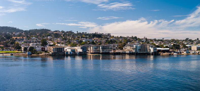 Monterey waterfront displays shoreline hotels, Cannery Row buildings, residential hills, and tranquil ocean bay waters under a bright sky along the California coast.