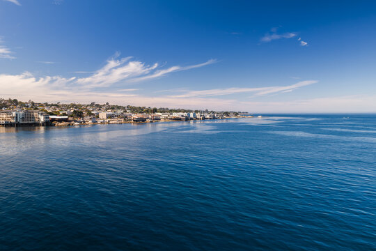 Aerial view shows the ocean water of Monterey Bay along the California shoreline with Cannery Row buildings under a sunny sky with wispy clouds during daytime.