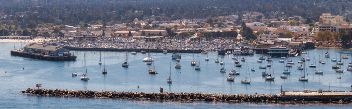 Aerial view of the coastal city of Monterey California showing sailboats anchored in the harbor alongside piers, breakwater rocks, and waterfront buildings.