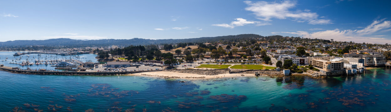 Aerial view presents the coastline of Monterey, California, showing Monterey Harbor, moored boats, coastal kelp beds, San Carlos Beach, and waterfront buildings on a sunny day.