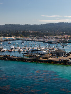 Aerial view shows boats moored along docks at Monterey Harbor in California, with the coastal shoreline, breakwater, and surrounding forested hills visible under daylight.