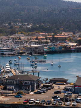 Aerial shot of Monterey harbor displaying moored sailboats, Fisherman's Wharf buildings, a parking lot, and forested hills under natural daylight.