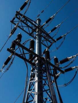 Large industrial electrical substation stands against clear blue sky background. High voltage pole system transmits significant amounts of power energy. Sturdy steel structure supports multiple