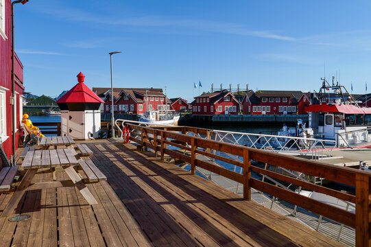 Sunny harbor scene in Svolvaer on the Lofoten Islands with a wooden terrace, boats and traditional red wooden houses beside the water under clear blue sky.