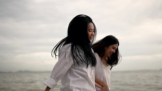 Two women walk side by side along a sandy shore in pale clothing. The two women pass ocean water under a cloudy sky, with palm trees lining the beach and open sand stretching along the coast.