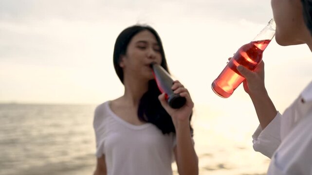 Two women hold beach drinks near the water, with glass bottles and clear cups of red liquid in their hands. The two women wear pale clothing on sand as the ocean and evening sky extend behind them.