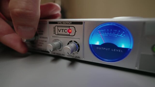 Close-up cropped shot of sound engineer hand adjusting output level knob on professional audio equipment. Blue VU meter showing increasing volume. Perfect for music production, studio content.