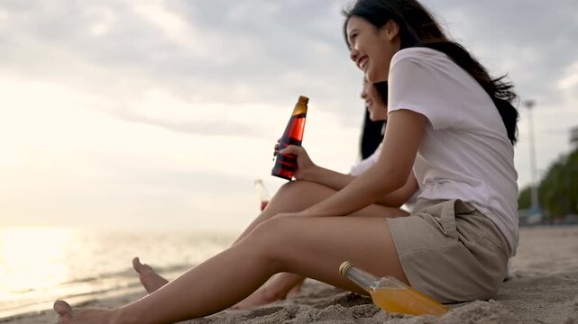 Two women sit together on a sandy shore with an ocean view and cloudy evening sky. The two women rest directly on the sand with bent or extended legs as small waves meet the beach in front of them.