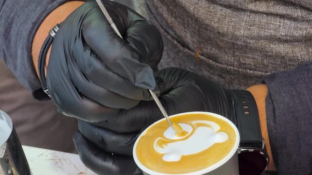 Close up of professional barista in black gloves drawing intricate latte art pattern on fresh coffee with stylus.