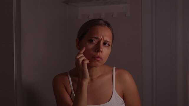 Young woman looking at face in bathroom mirror and checking small pimple, acne, redness and skin imperfections, touching cheek with worried expression, skincare concern