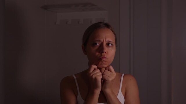 Young woman pinching jowls in mirror with dissatisfied expression, concerned about facial aging, skin sagging and loss of firmness