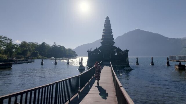 Tourist woman walking on a wooden bridge towards Pura Ulun Danu Bratan water temple on Lake Bratan, Bali, Indonesia.