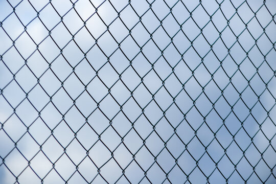 Chain-link fence silhouetted against a pale blue cloudy sky, creating a clean geometric pattern with atmospheric depth and a minimalist outdoor composition.
