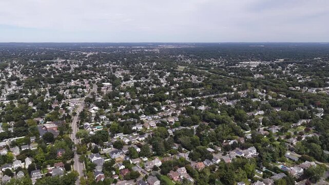 A drone glides over leafy residential neighborhoods in Babylon on Long Island in New York. Detached houses and winding streets stretch toward the horizon under a light sky with dense green tree cover.