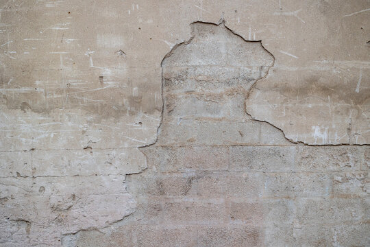 Close-up of an aged plaster wall with a large peeling section, visible cracks, exposed layers, faded beige tones, and authentic architectural deterioration.