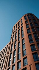 Fototapeta premium Modern red brick apartment building stands under clear blue sky. Contemporary residential architecture features curved facade with many windows, balconies. Urban housing structure shows geometric