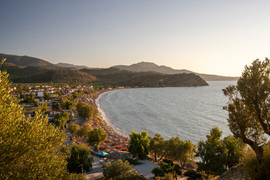 Panoramic view across Anaxos Bay on Lesbos, Greece, with its sandy beach, olive trees, seaside village and rugged mountains glowing in warm evening light.