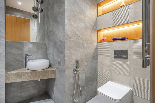 A room with gray tiles, a wash basin on a stone counter, and a toilet. Warm light from the shelves highlights the modern design. Water fixtures are