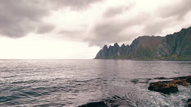 Scenic drone shot moving backward over a rocky coastal shore with dramatic mountain peaks and ocean waves in Norway