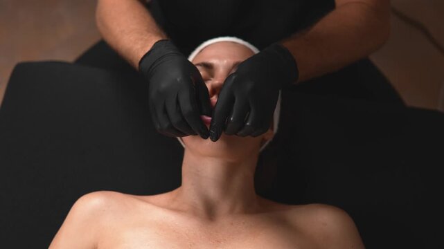 In a wellness session, a massage therapist uses gloved hands to massage a relaxed woman's face in a spa setting. The front close view highlights beauty care, client comfort and client care.