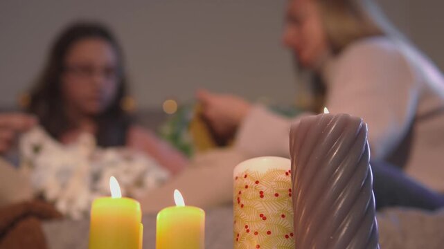 Friends craft a popcorn garland behind softly glowing foreground candles during the holiday season. Shallow focus view presents cozy creative evening with strong commercial appeal.