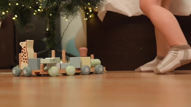 A toddler boy stands beside a wooden train while exploring a festive play area in a festive home. The low feet view conveys gross motor skills, child development and family warmth.