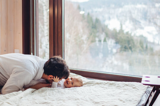 Happy family playing on bed enjoying snowy mountains landscape from hotel window. Dad having fun with toddler baby girl