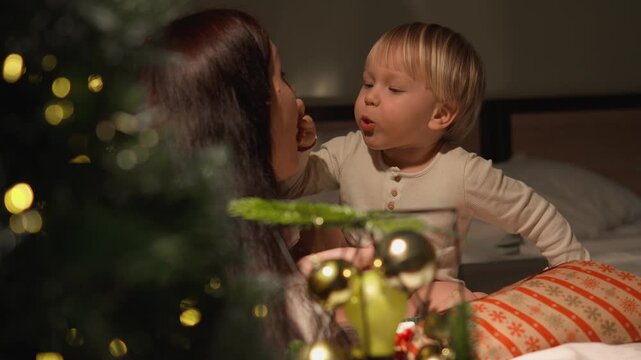 A toddler boy examines a decorated lantern while his mother watches in a festive home. The close profile view conveys guided discovery, family learning and family warmth.