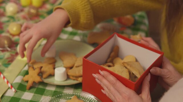 Detailed holiday footage captures hands as they place cookies and marshmallows inside a red Christmas gift box. Overhead detail view presents homemade present making with strong commercial appeal.