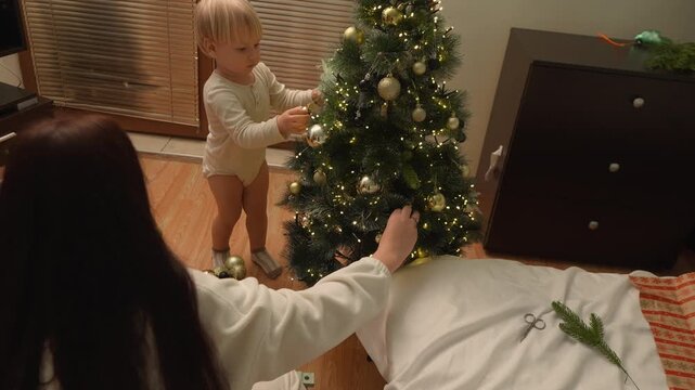 A toddler boy places an ornament on the lower branches with his mother watching in a festive home. The wide floor view conveys toddler independence, Christmas tradition and family warmth.
